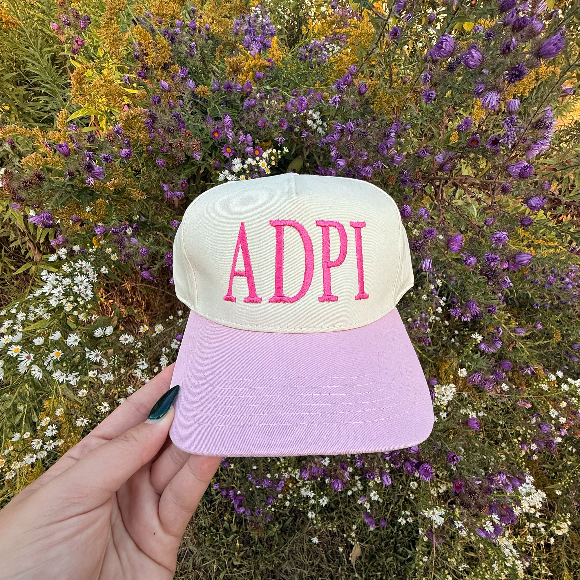 A person's hand holding a white baseball cap with pink letters against a background of colorful wildflowers.