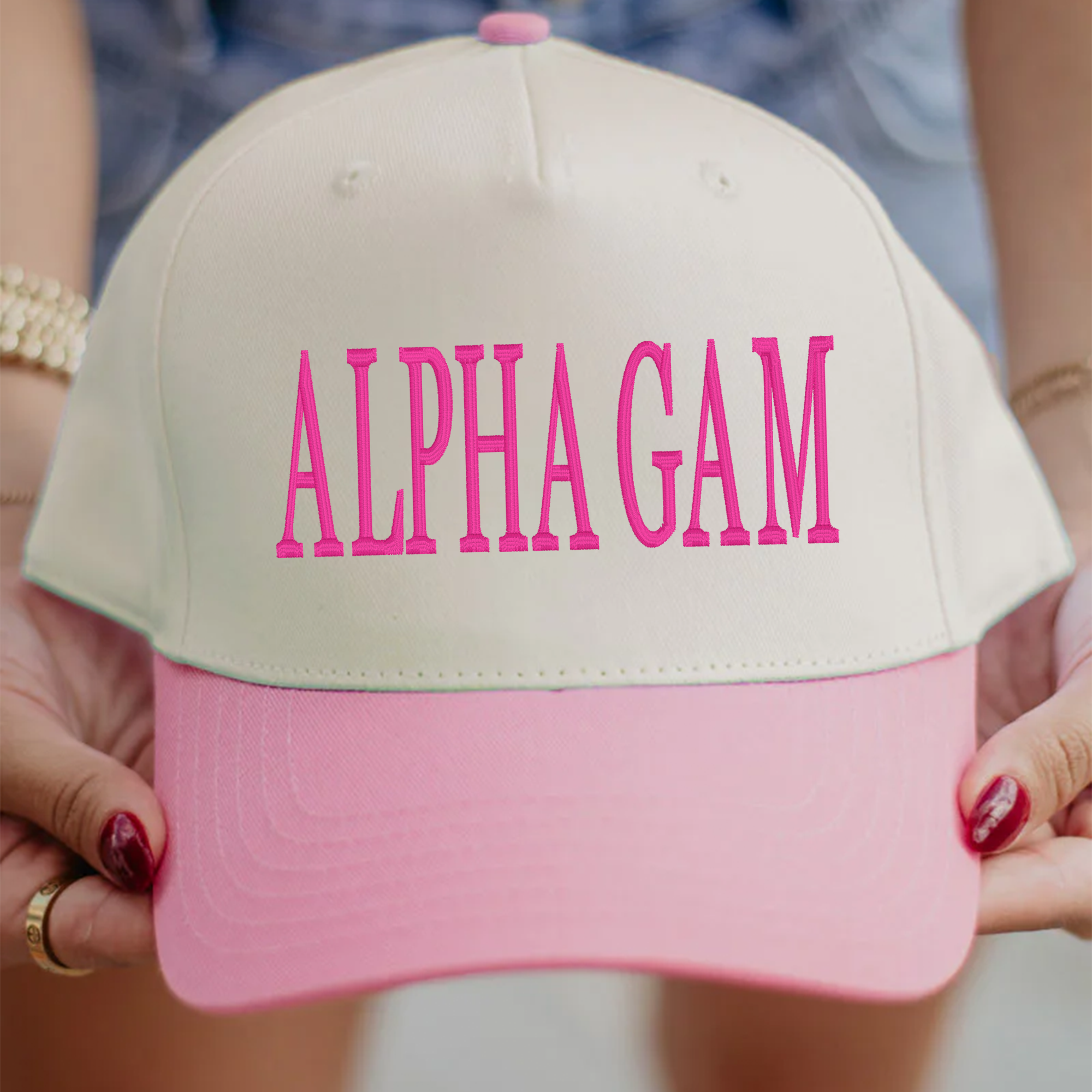 A person is holding a pink and white baseball cap with the text "ALPHA GAM" printed on it.