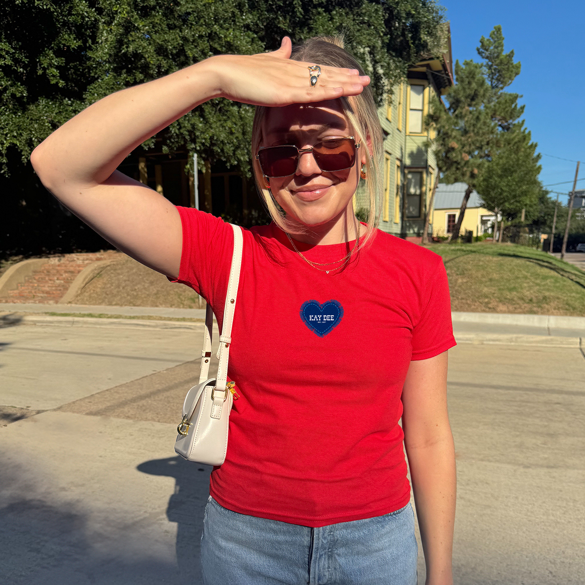 A woman wearing a red t-shirt with a heart design and sunglasses stands on a street, with a hand covering her face.