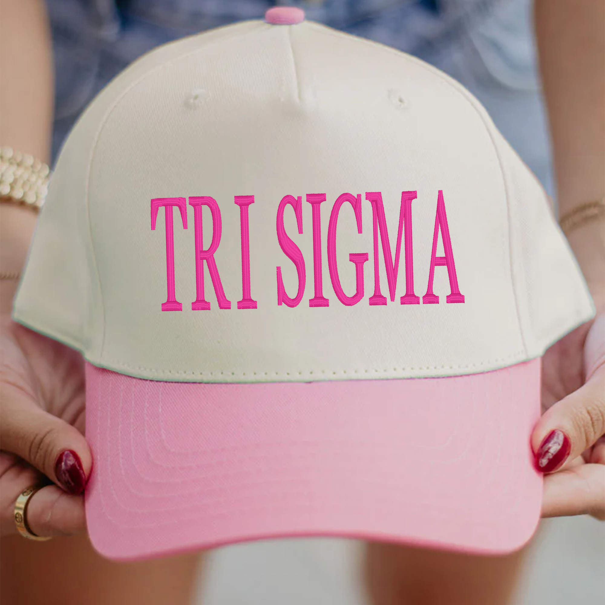 A person is holding a white and pink baseball cap with the text "TRI SIGMA" printed on it.