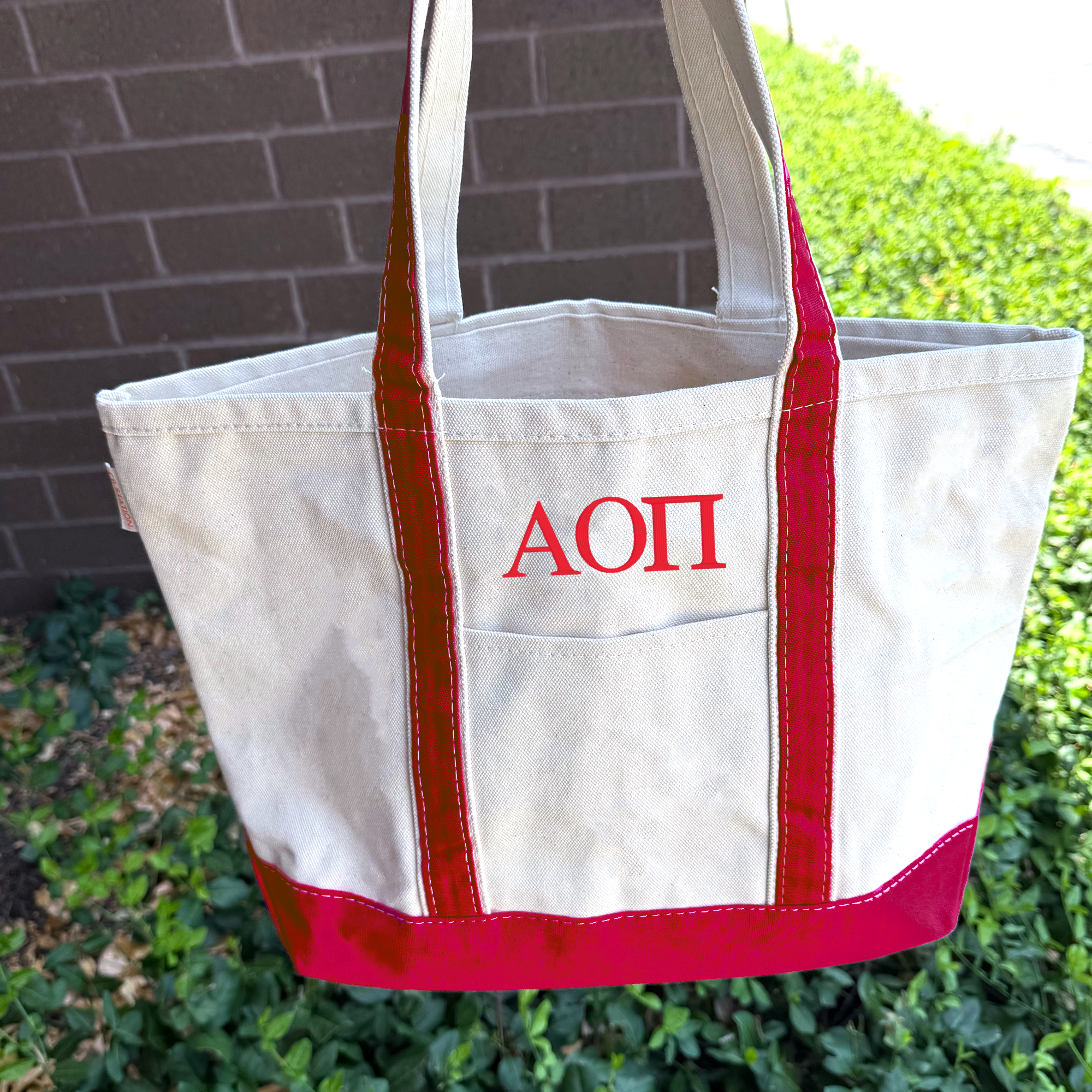 A white tote bag with red trim and the Greek lettering "ΑΓΟΡΕΙΚΗ" printed on it, hanging on a brick wall.
