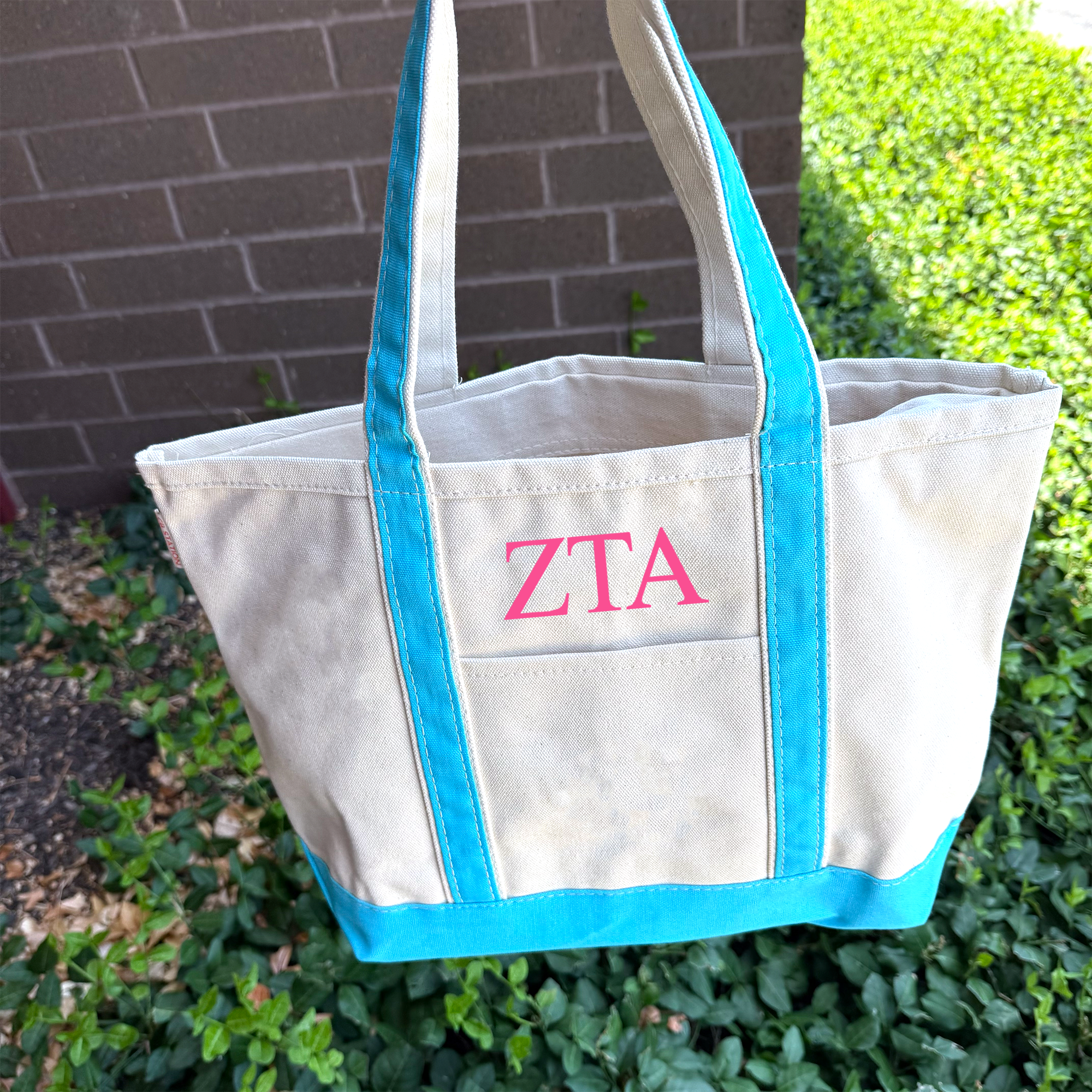 A white tote bag with blue trim and the letters "ZTA" printed on it, hanging on a brick wall.