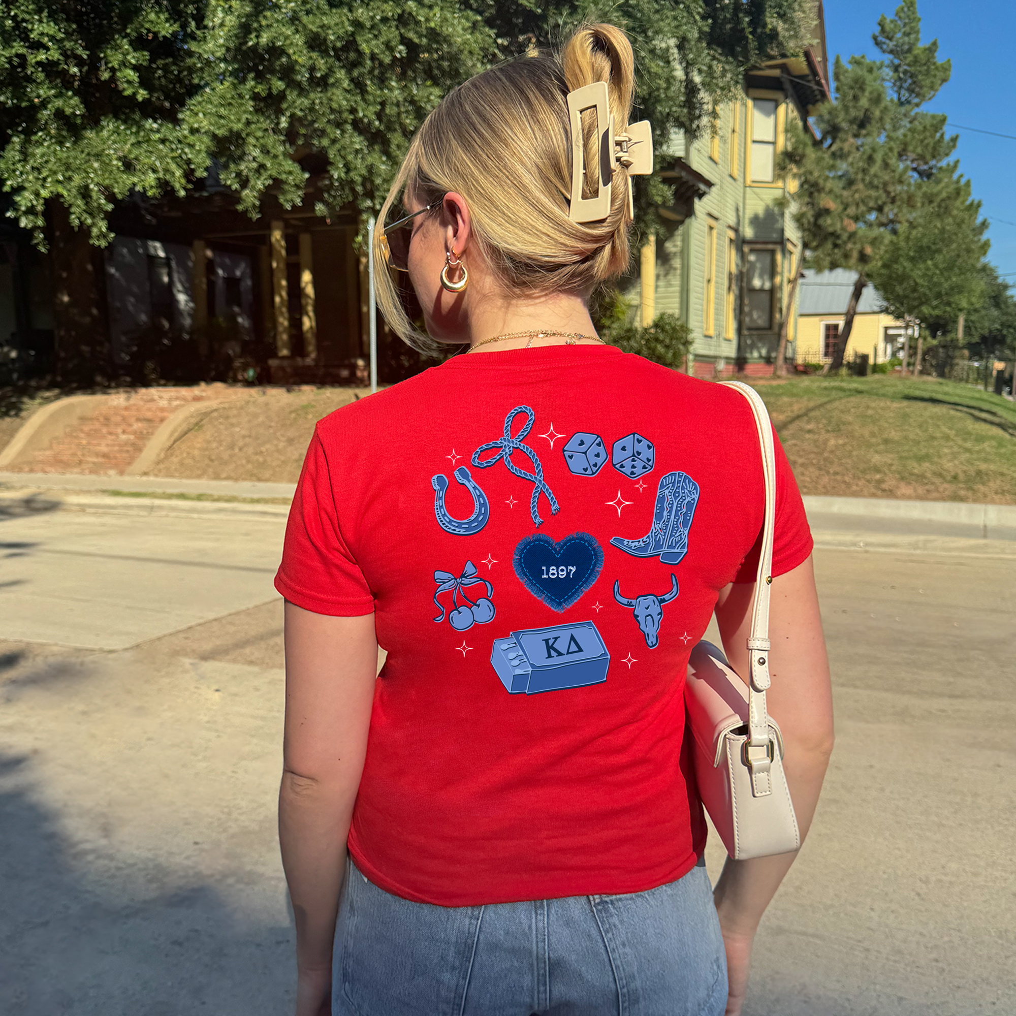 A woman wearing a red t-shirt with various icons and text on the back is standing on a street.