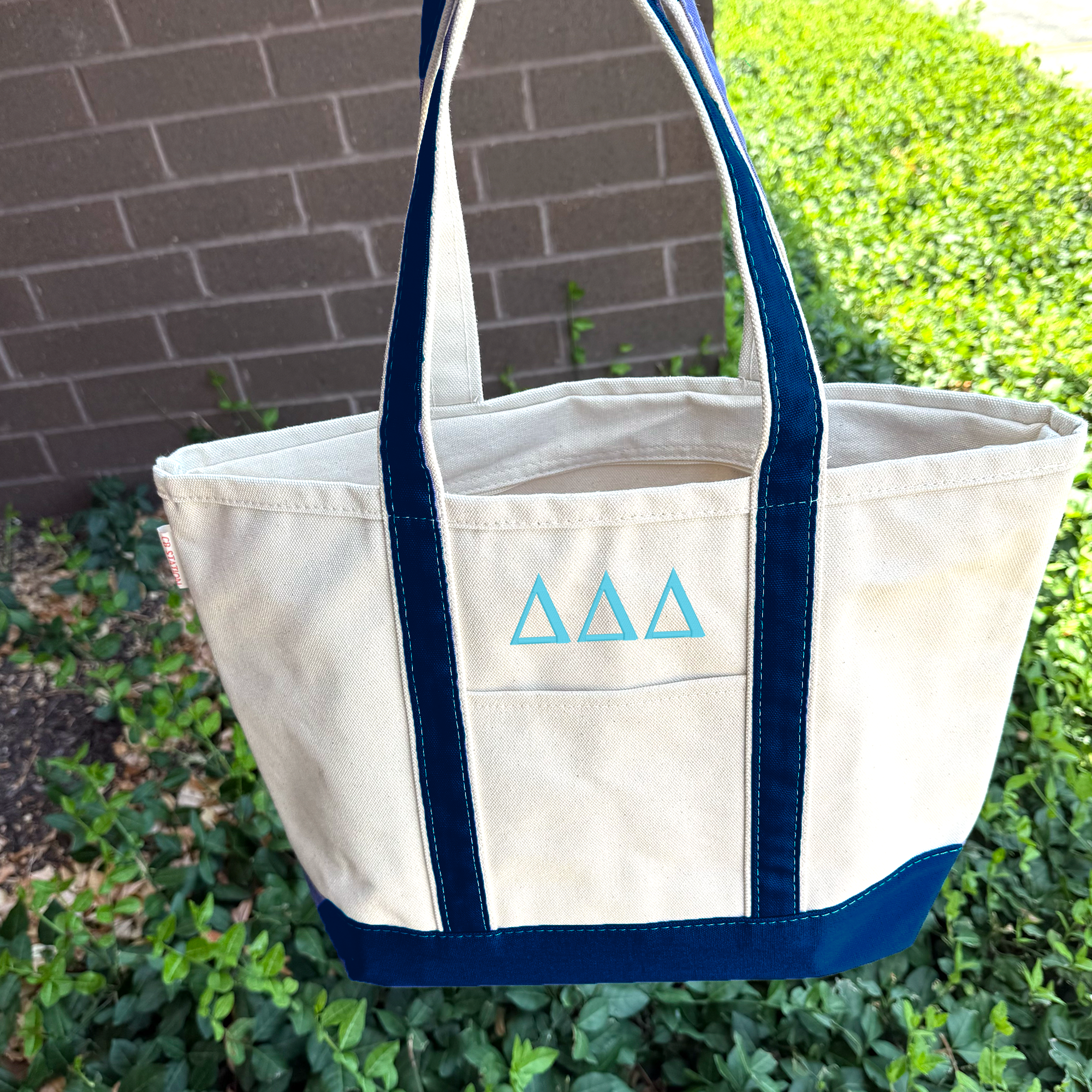 A white tote bag with blue trim and three blue triangles on the front, hanging on a grassy area with a brick wall in the background.