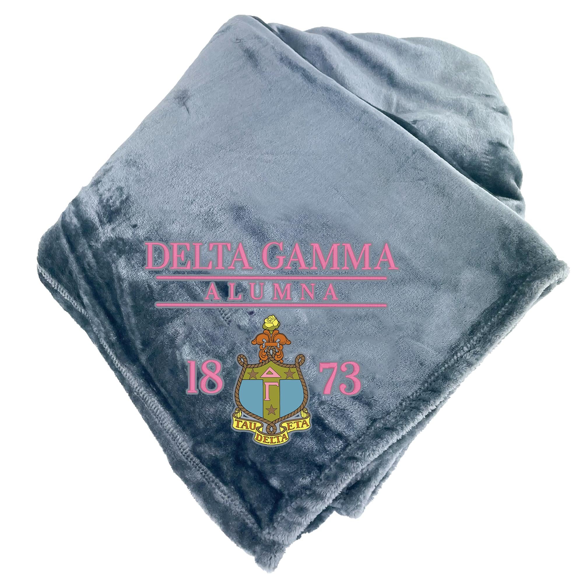 A gray fleece blanket with the text "DELTA GAMMA ALUMNA 1873" printed on it, along with a coat of arms.