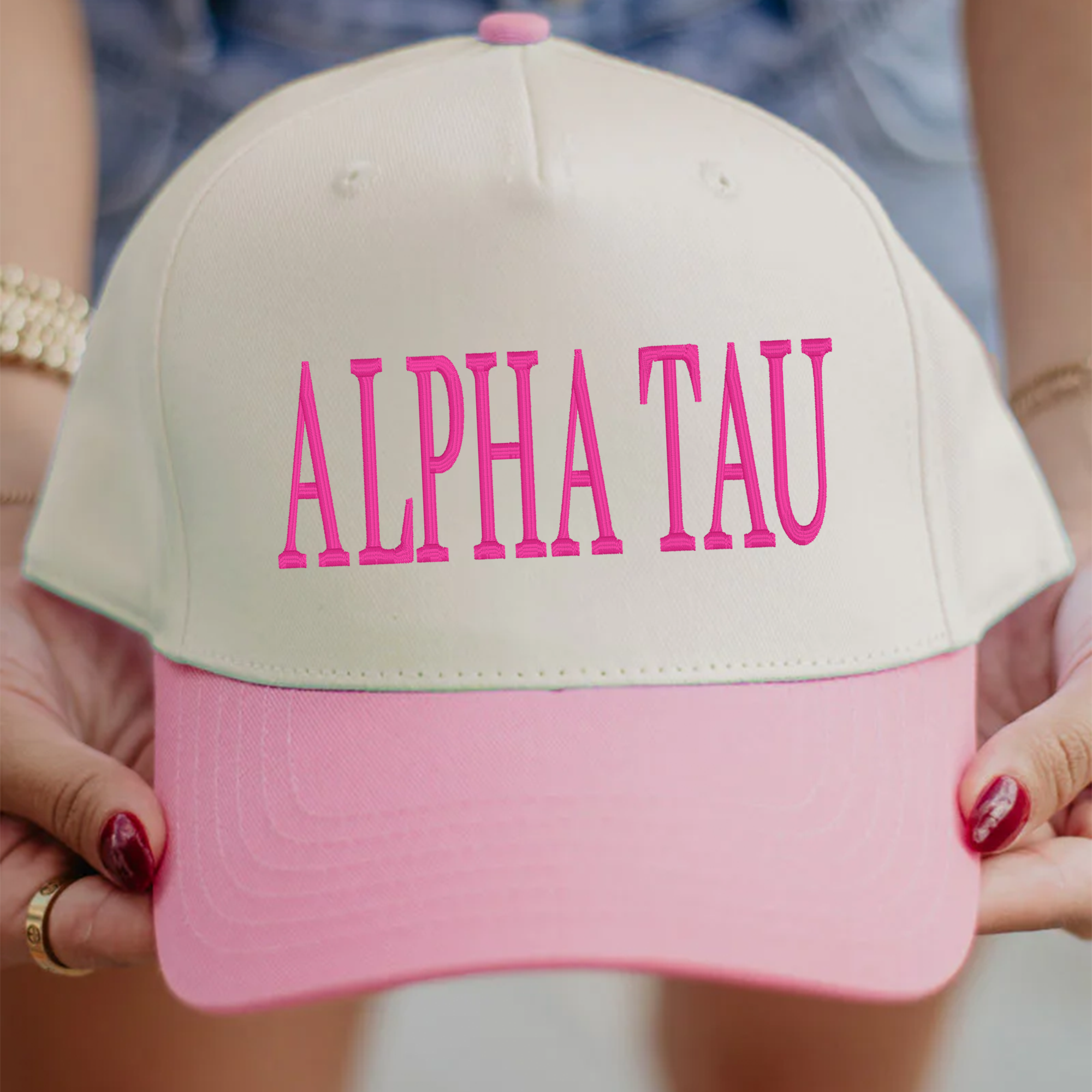 A person is holding a pink and white baseball cap with the text "ALPHA TAU" printed on it.