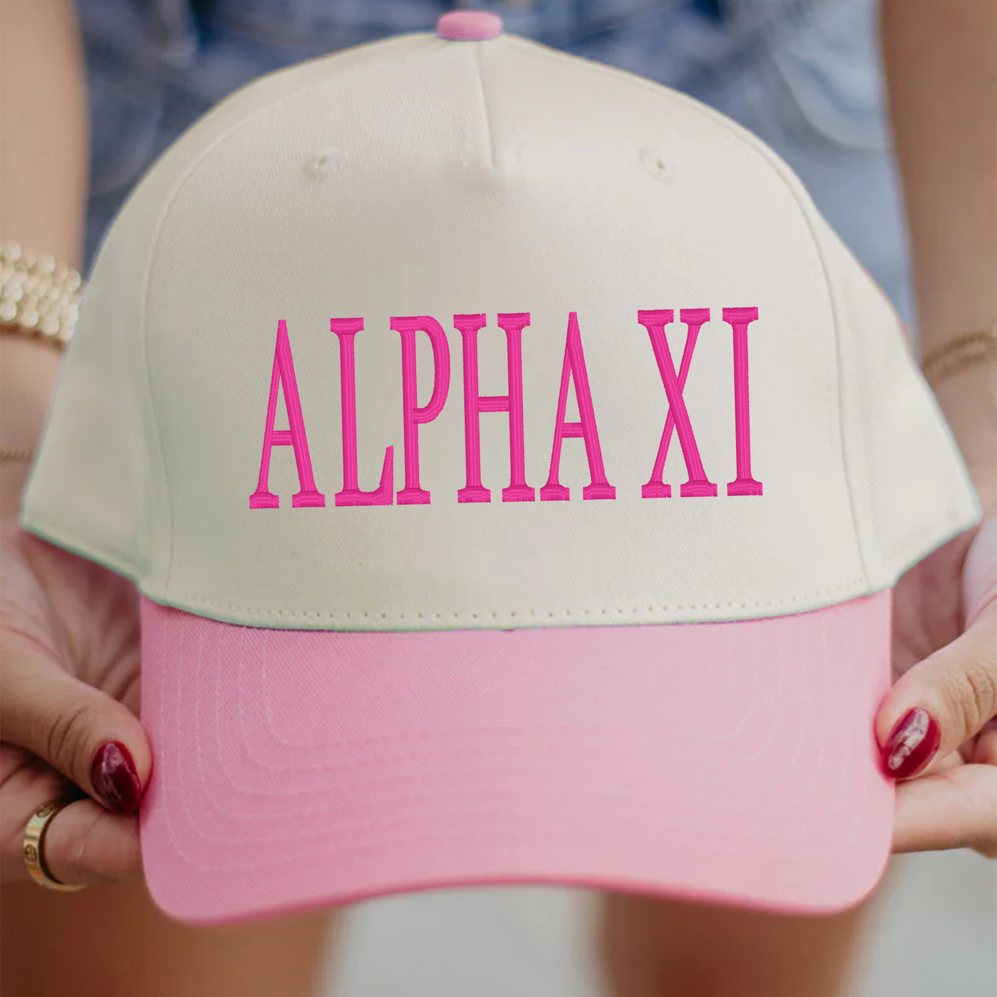 A person is holding a white and pink baseball cap with the text "ALPHA XI" printed on it.