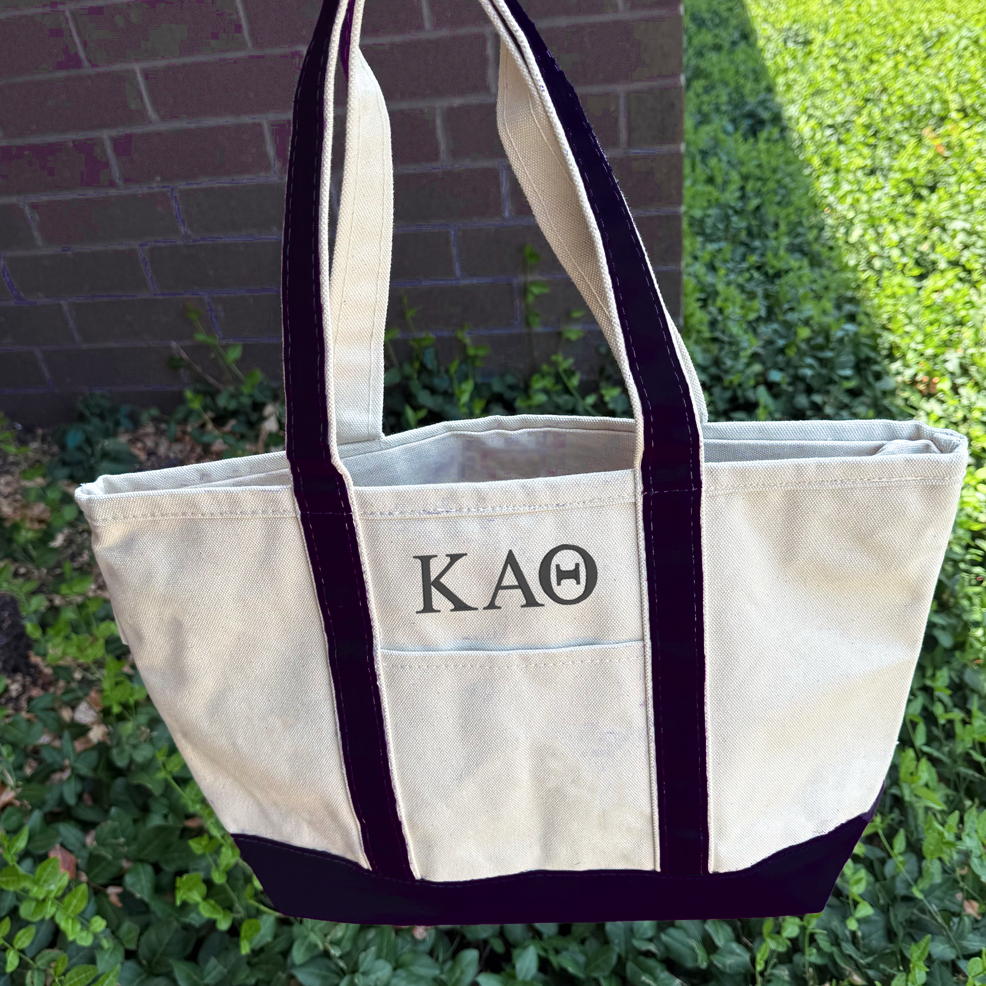 A white tote bag with black straps and the letters "KA" printed on it, placed on a grassy area next to a brick wall.