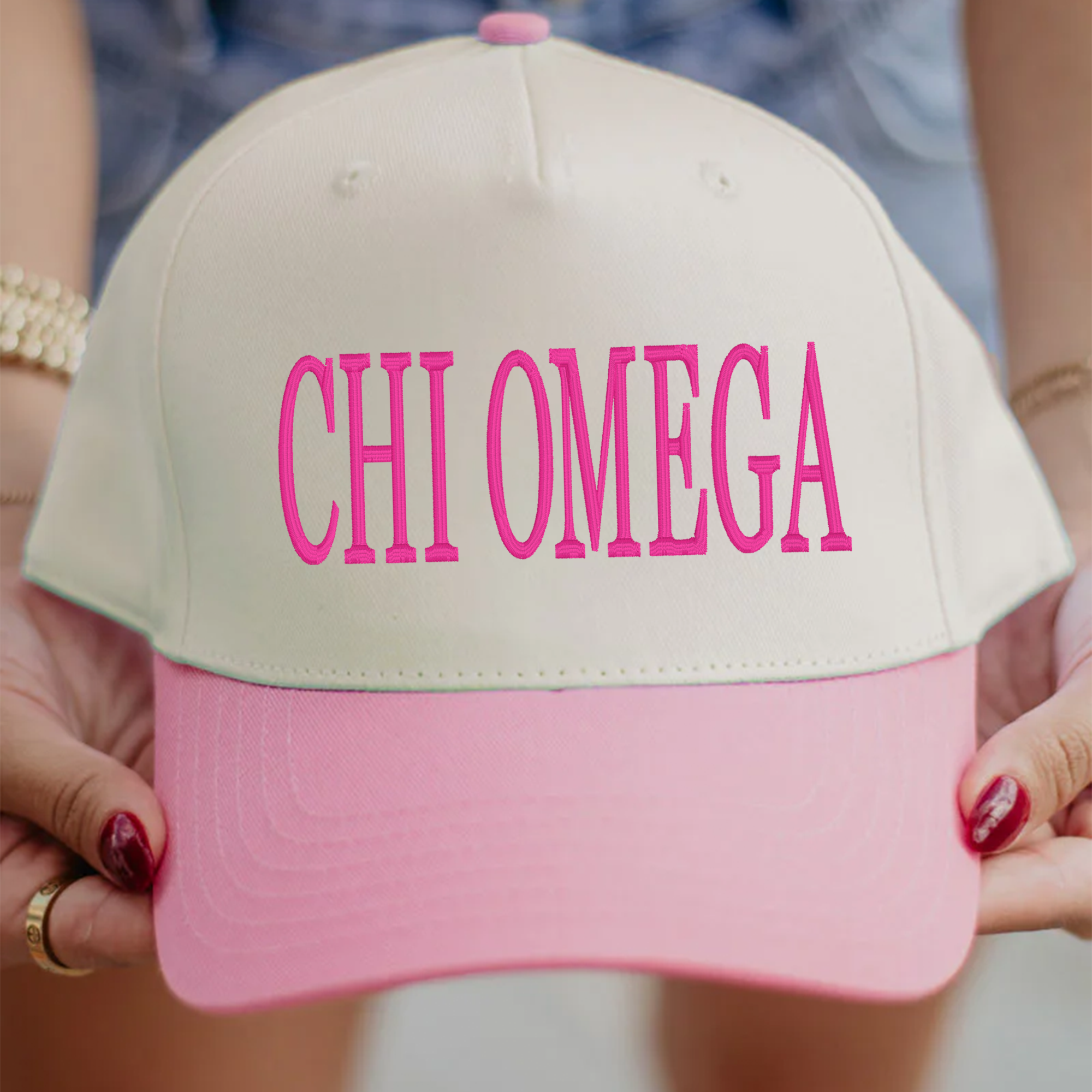 A person is holding a pink and white baseball cap with the text "CHI OMEGA" printed on it.