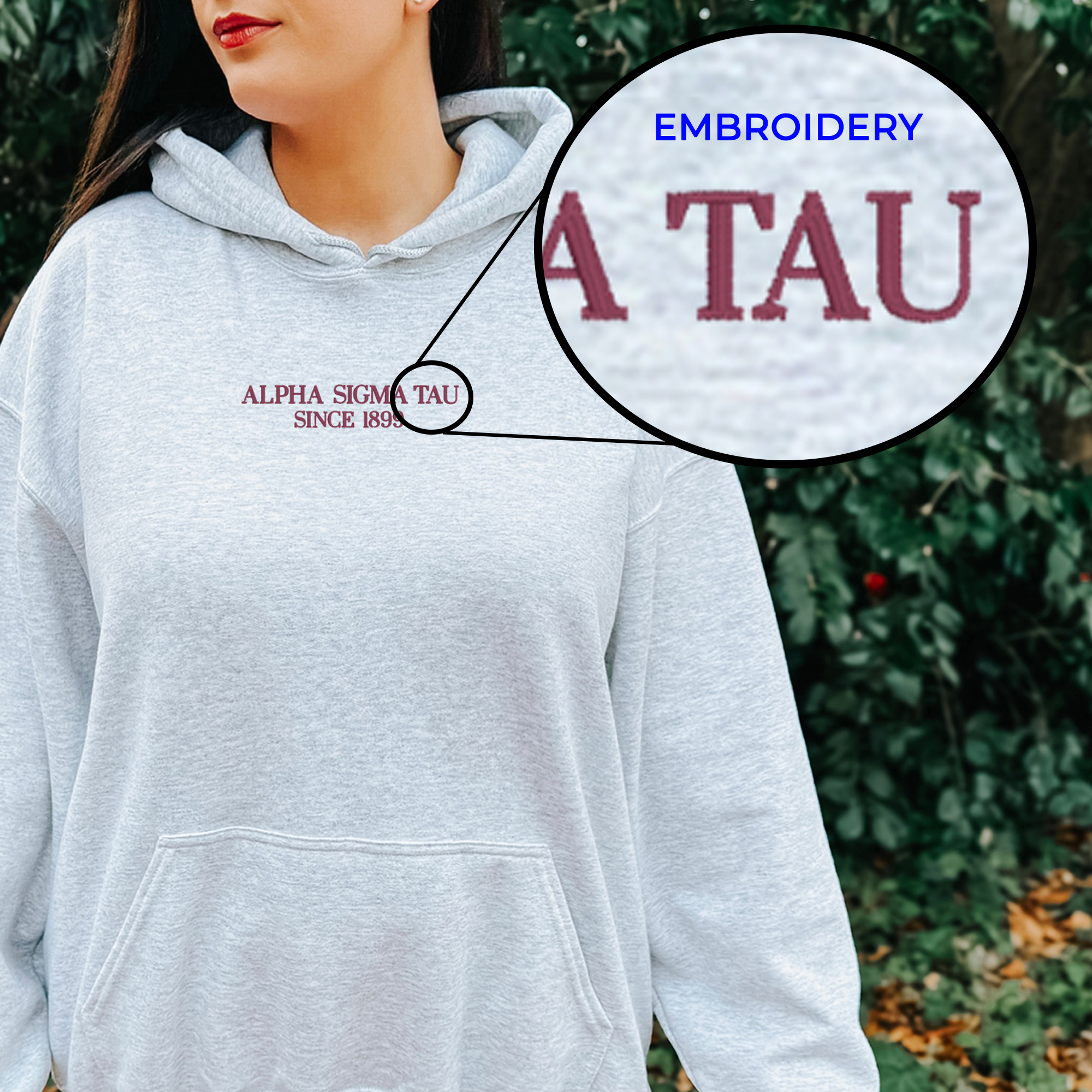 A woman wearing a grey hoodie with the embroidered text "ALPA SIGMA TAU SINCE 1898" on it, standing in front of a background of greenery.