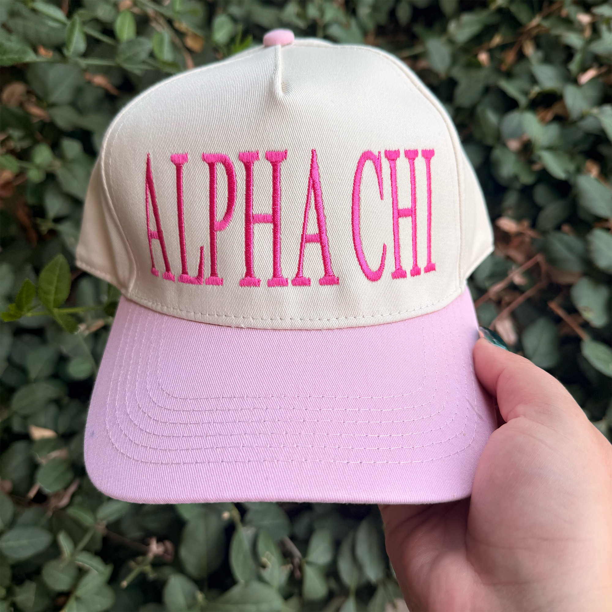 A person's hand is holding a pink and white baseball cap with the text "ALPA CHI" embroidered on it.