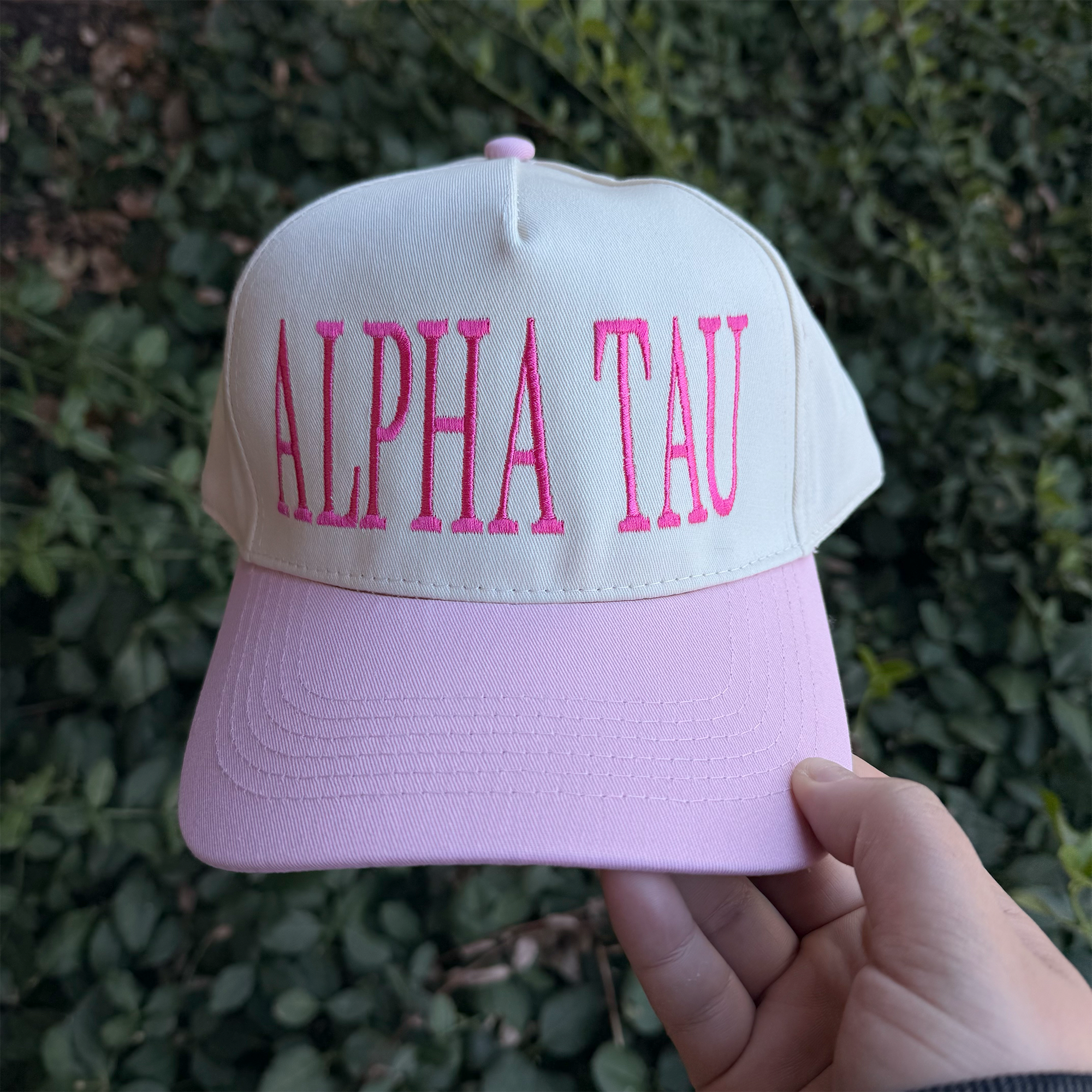 A person is holding a white baseball cap with pink text on it, which says "ALPHA TAU".