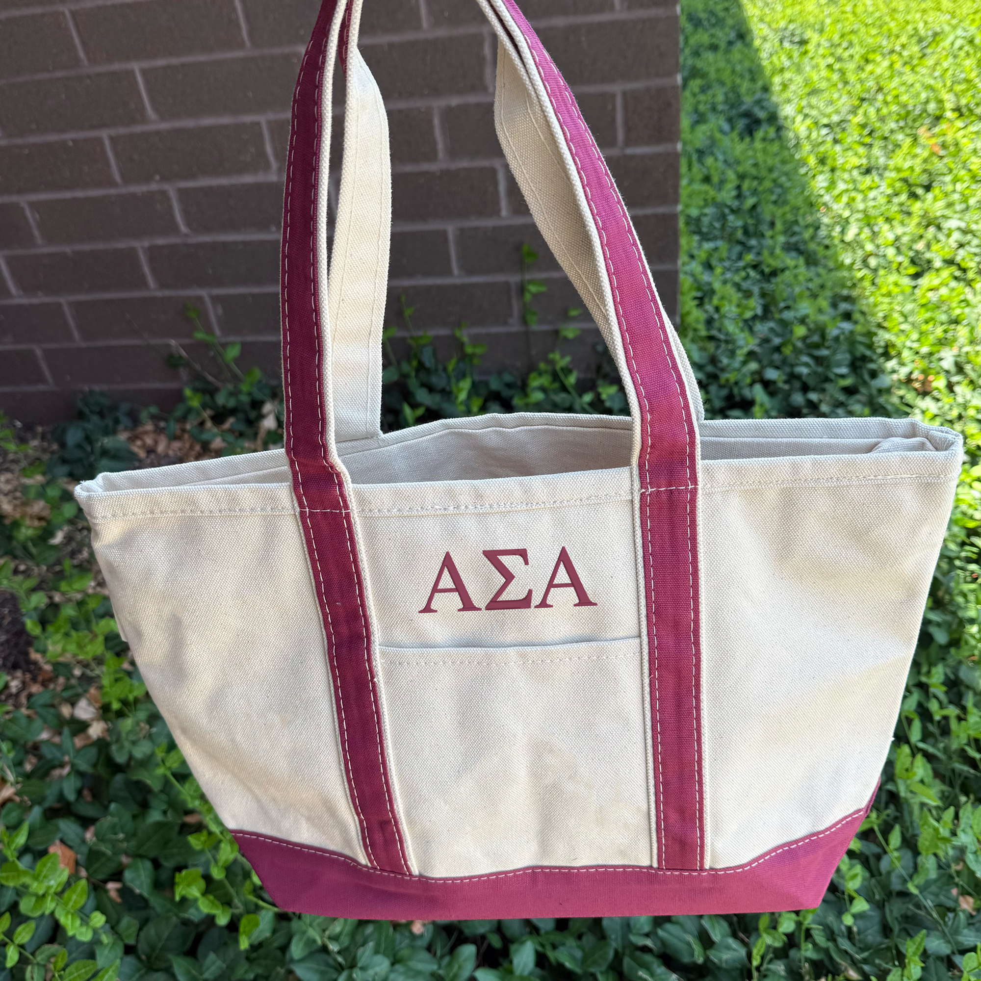 A beige tote bag with maroon trim, featuring the initials 'ΑΕΛΑ' printed on it, is displayed against a brick wall and surrounded by greenery.