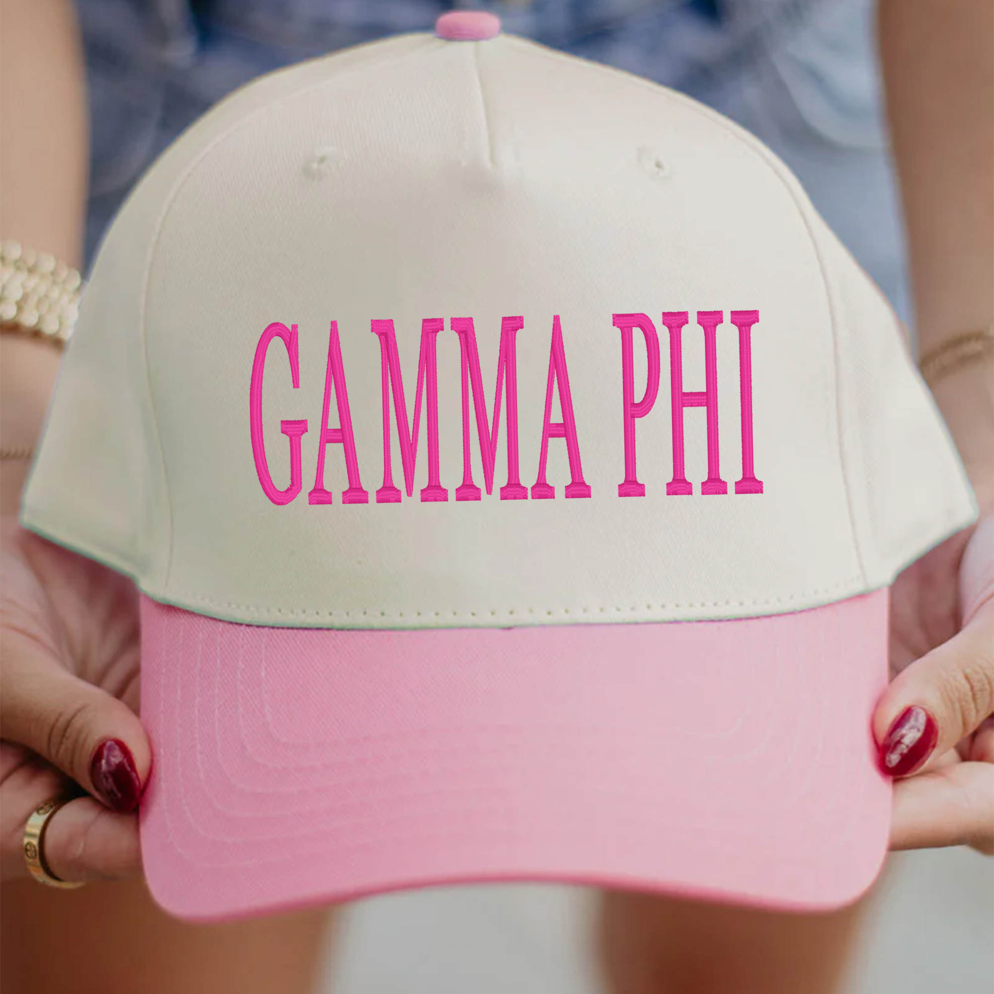 A person is holding a white and pink baseball cap with the text "Gamma Phi" printed on it.