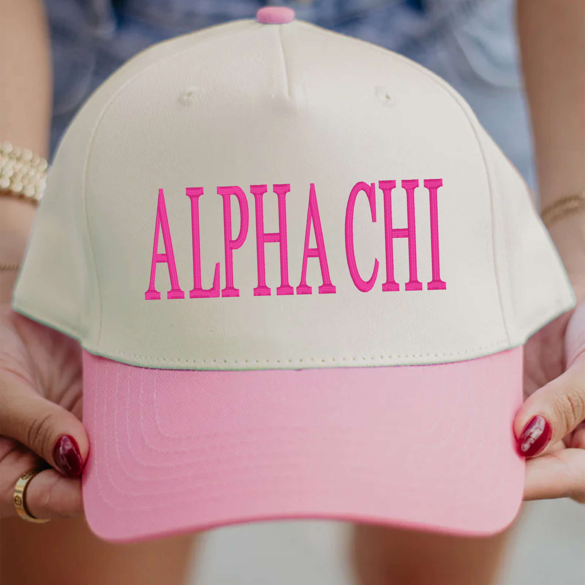 A person is holding a pink and white baseball cap with the text "ALPHA CHI" printed on it.