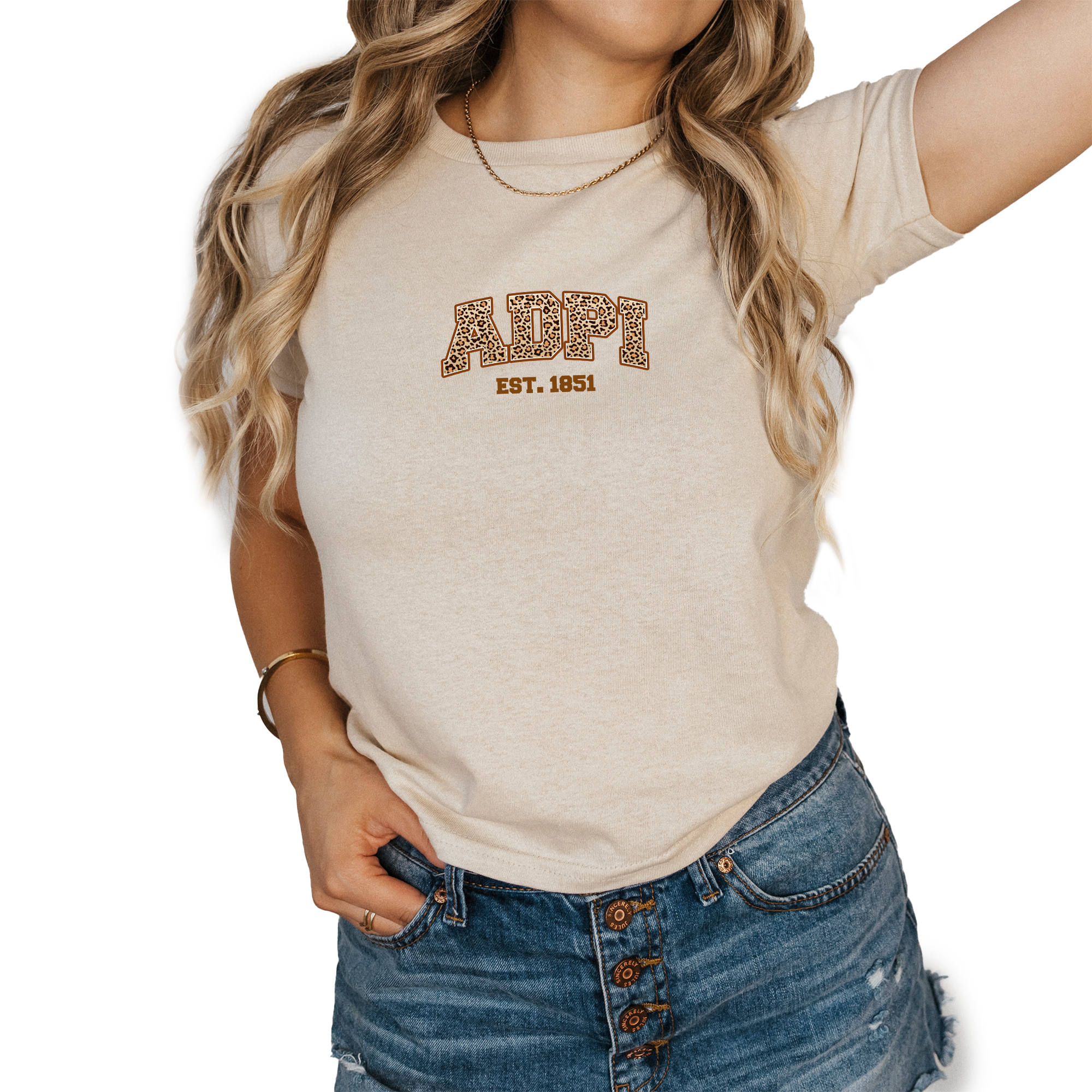 A woman wearing a beige t-shirt with the text "ALPI EST. 1851" printed on it, along with a chain necklace. She is also wearing blue denim shorts.